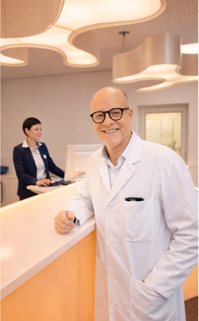 Lächelnder Arzt mit weißem Kittel und Brille an einer hellen Empfangstheke, im Hintergrund eine Frau, die an einem Computer arbeitet.