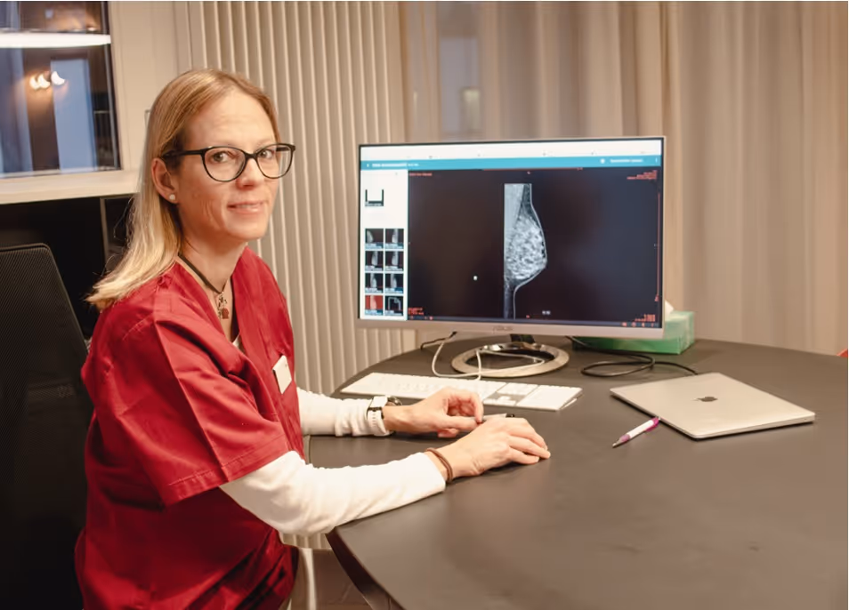 Frau in roter medizinischer Arbeitskleidung sitzt an einem Schreibtisch vor einem Monitor mit einer Mammographieaufnahme.