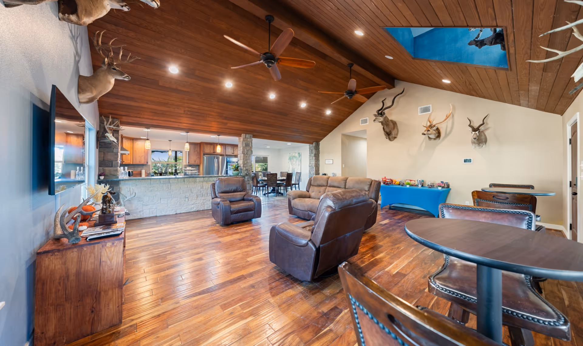 Spacious living room with leather armchairs and sofa, wood-paneled slanted ceiling with fans, mounted deer heads on wall, and kitchen in background.