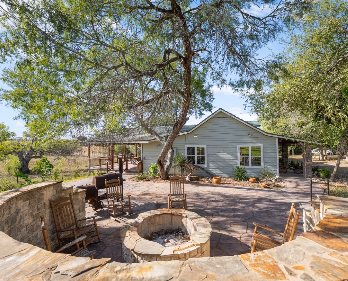 Lodge patio with stone fire pit surrounded by wooden rocking chairs and a light gray house with trees in the background.