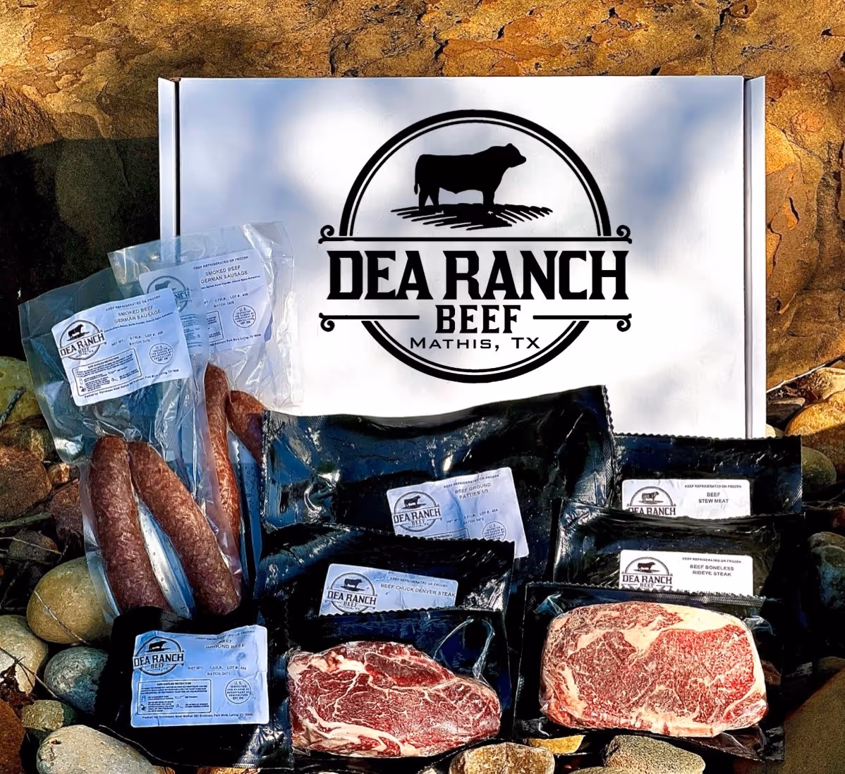 Box of DEA Ranch Beef from Mathis, TX, surrounded by various vacuum-sealed cuts of beef and smoked sausages displayed on rocks.