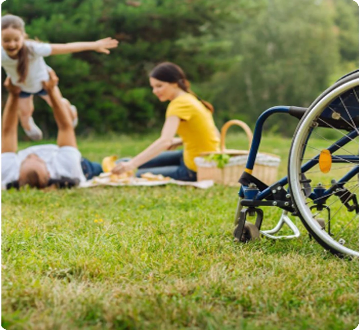 Un homme allongé sur l'herbe soulève joyeusement une petite fille en l'air tandis qu'une femme assise à côté d'eux prend des aliments sur une couverture de pique-nique près d'un fauteuil roulant.