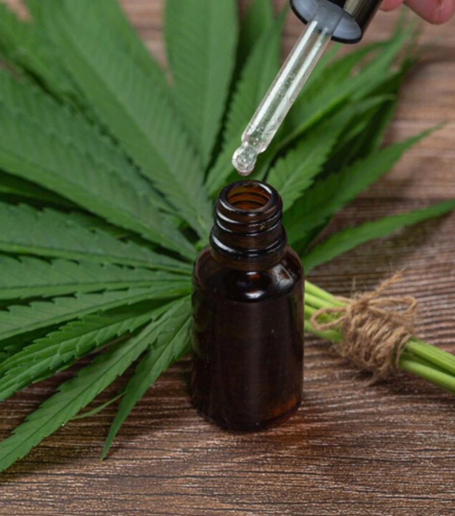 Goutte d'huile tombant d'un compte-gouttes dans un flacon en verre devant une feuille de cannabis sur une surface en bois.