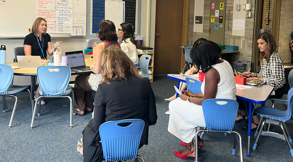 A group of six educators engaged in a meeting inside a classroom, with some seated at tables and others on blue chairs, laptops and notebooks in use, and whiteboards with notes in the background.