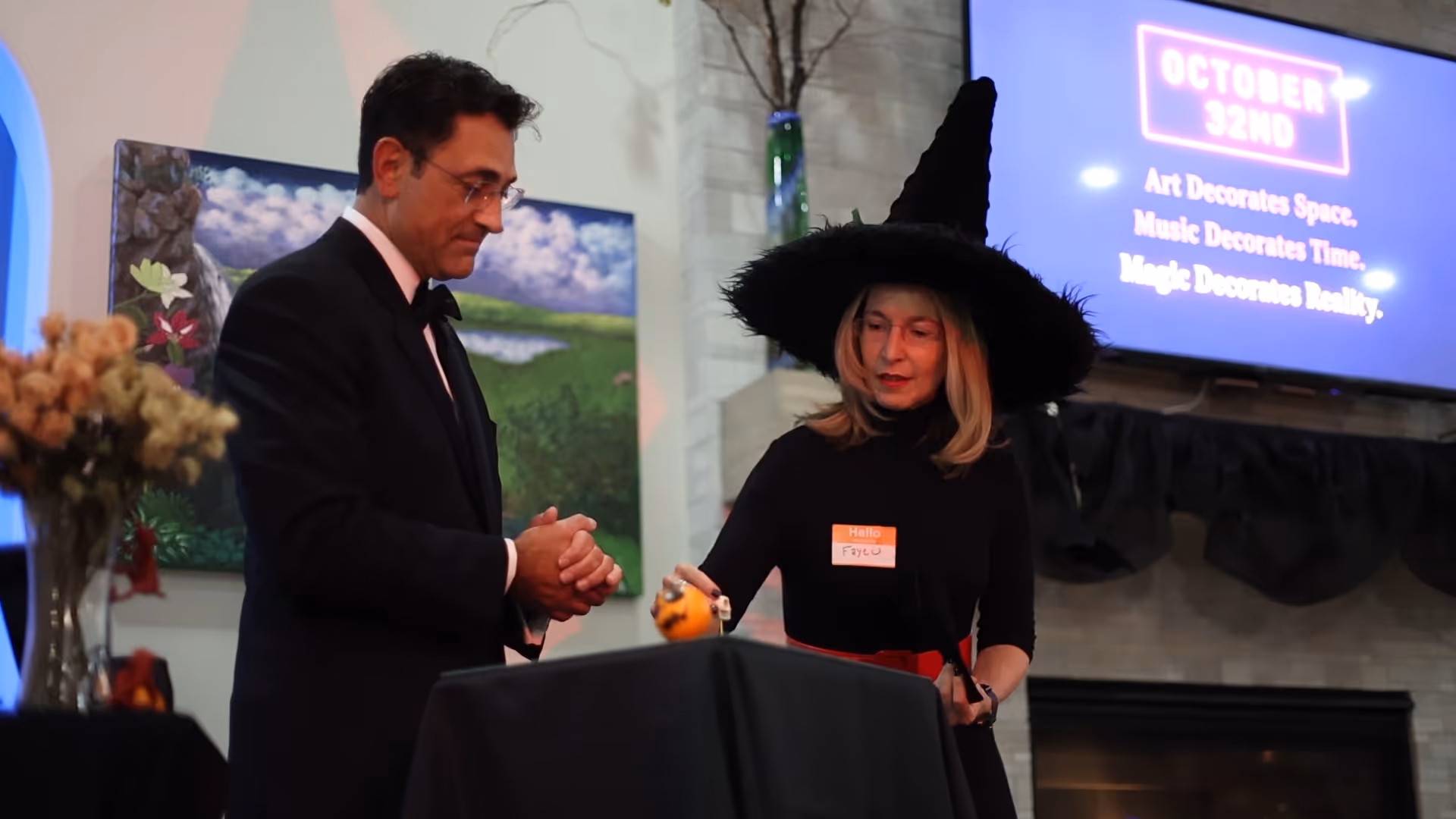 Man in tuxedo and woman dressed as a witch with a pointed hat standing by a table with Halloween decorations.