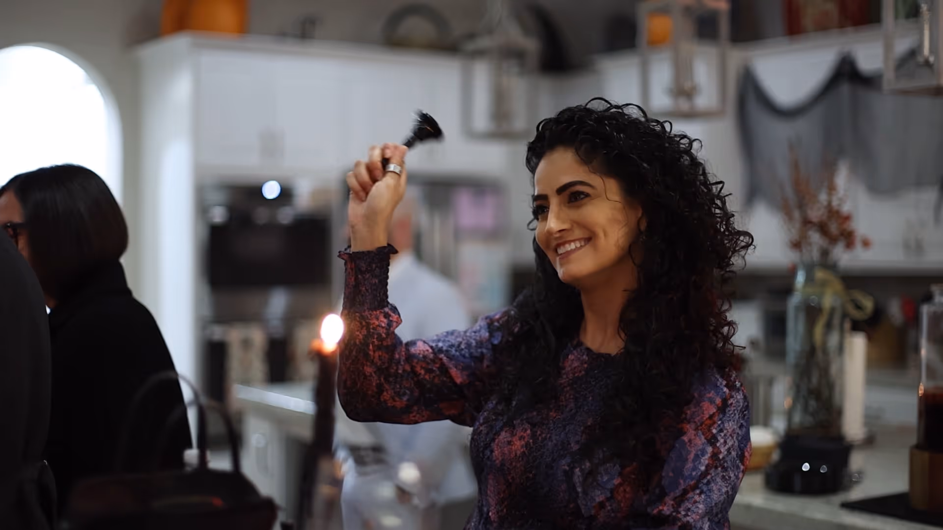 Smiling woman with curly hair ringing a small bell in a kitchen setting with blurred people and candle in the background.