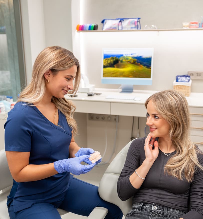 Zdravotní sestra v modrém uniformě s rukavicemi drží dentální model a mluví s usmívající se pacientkou v ordinaci.
