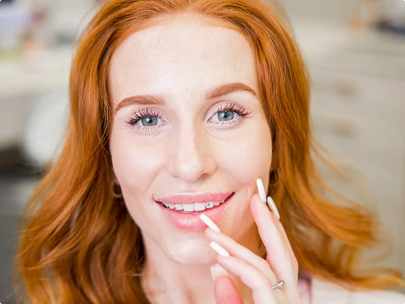Usměvavá žena s rezavými vlasy a světle modrýma očima, která si jemně dotýká tváře s dlouhými bílými nehty.