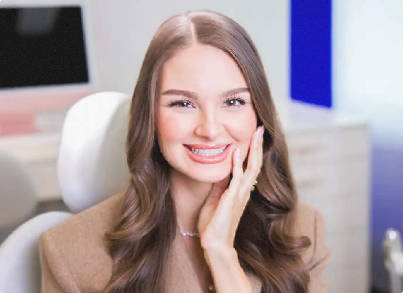 Mladá žena s krásnými vlnitými vlasy a rozzářeným úsměvem s rovnými zuby, která si lehce sahá na tvář.