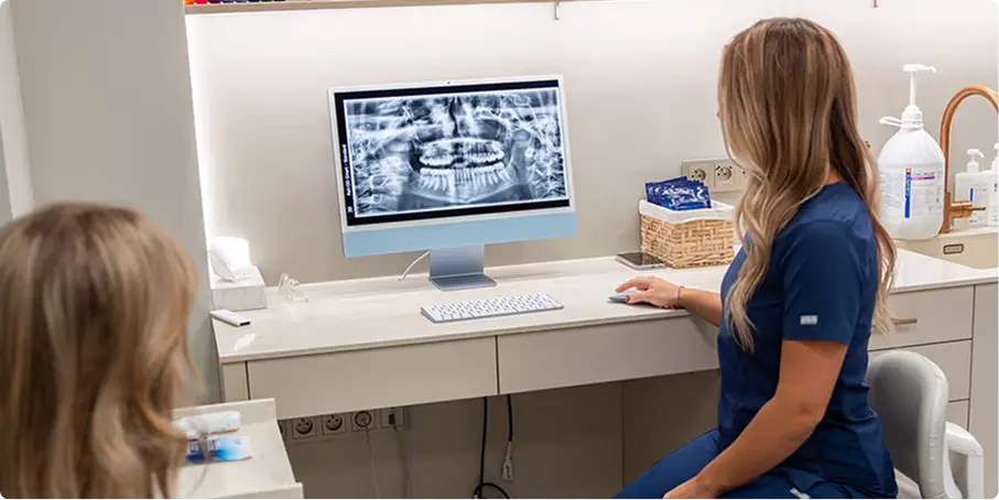 Žena v lékařském oblečení sedí u stolu a ukazuje pacientce rentgen zubů na počítači v zubní ordinaci.