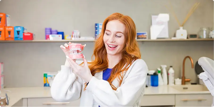 Žena v lékařském plášti v zubní ordinaci drží a ukazuje model lidských zubů a dásní.