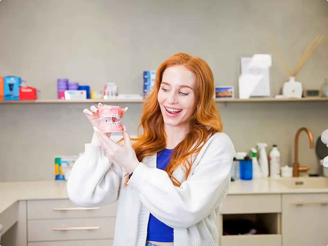 Usměvavá žena s rezavými vlasy drží dentální model se zubními rovnátky v zubní ordinaci.