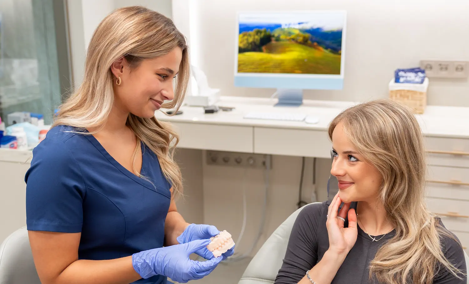 Dentální hygienistka v modré uniformě drží model zubů a ukazuje jej pacientce sedící na zubním křesle.