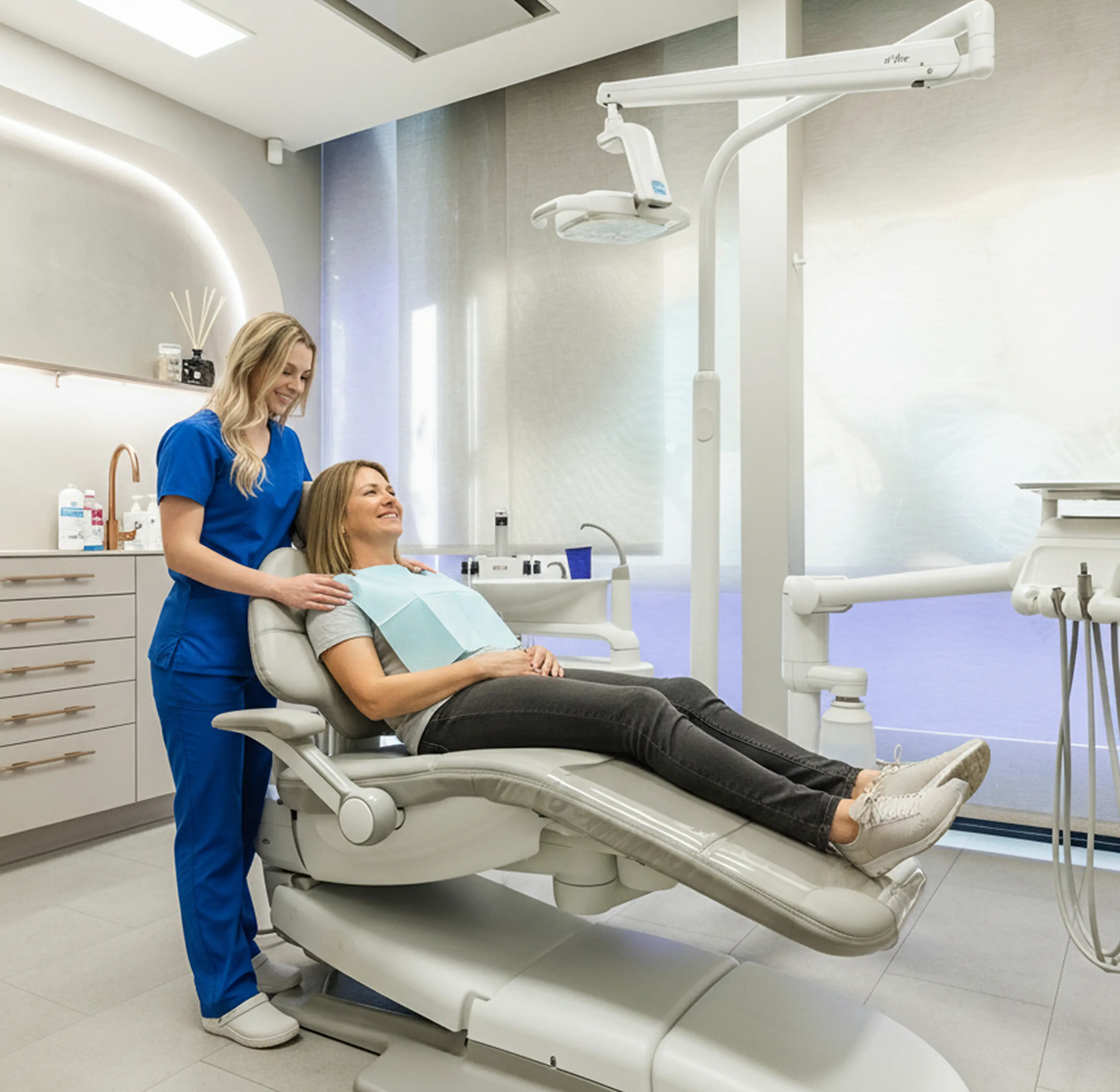 Pacientka sedící v zubním křesle s modrým pláštěm a usměvavá ošetřující zubní hygienistka v modré uniformě v moderní zubní ordinaci.