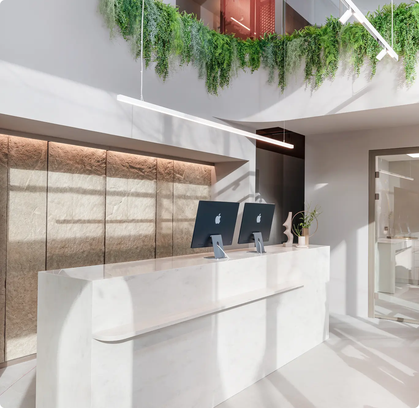 Recepční pult ordinace MILES na Praze 5 Andělu, z bílého mramoru se dvěma počítači Apple v moderním prostorově světlém interiéru s visícími zelenými rostlinami.