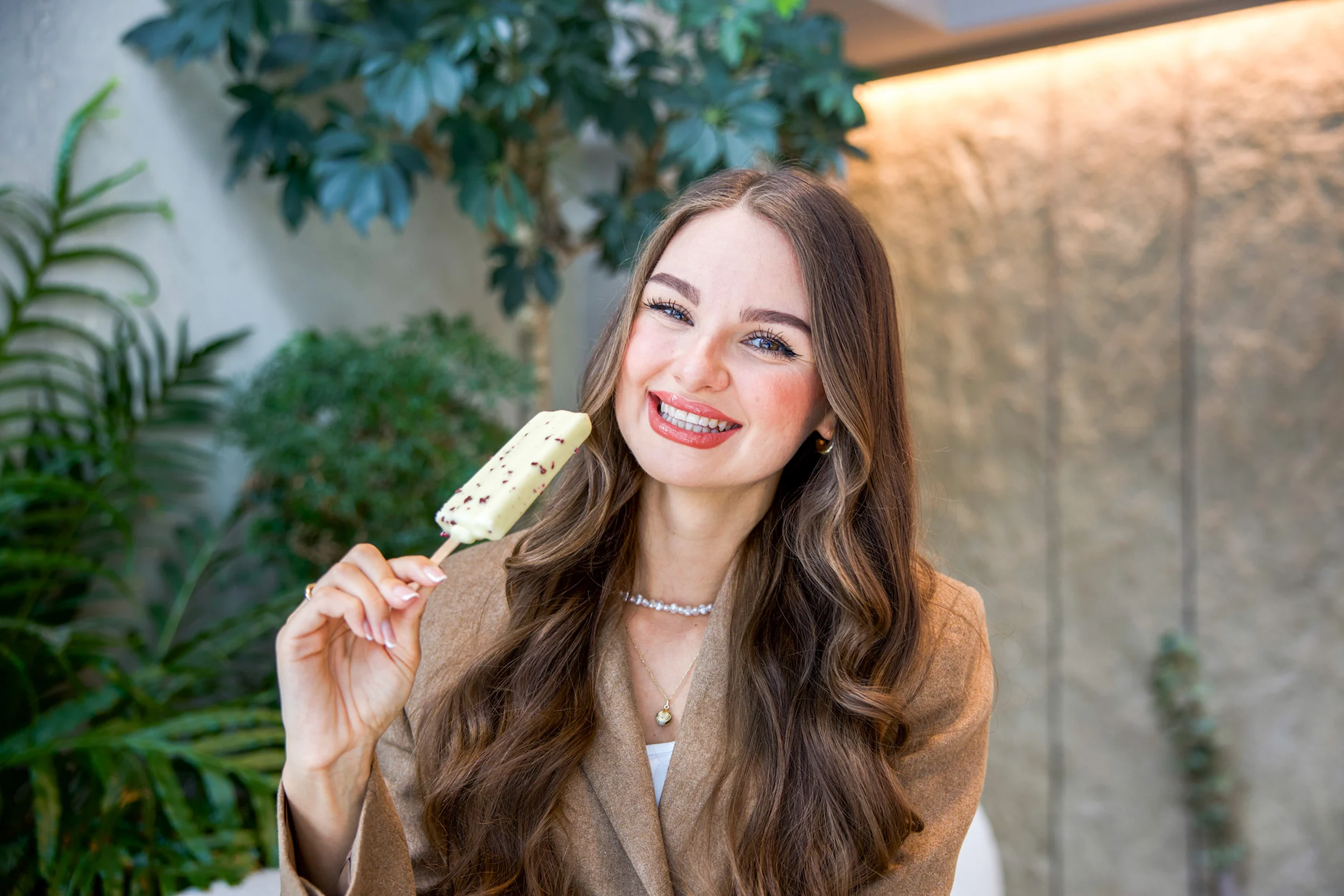 Usměvavá žena s dlouhými vlnitými vlasy drží nanuk v interiérovém prostředí s rostlinami.