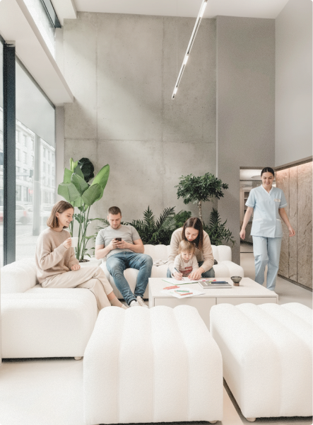 Rodina sedí v moderní světlé čekárně s bílými pohovkami, rostlinami a zdravotní sestrou přicházející z chodby.