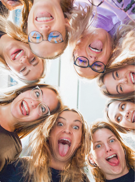 Skupina sedmi veselých zaměstnankyň kliniky MILES dívajících se dolů s úsměvy a jazykem venku v kruhu.