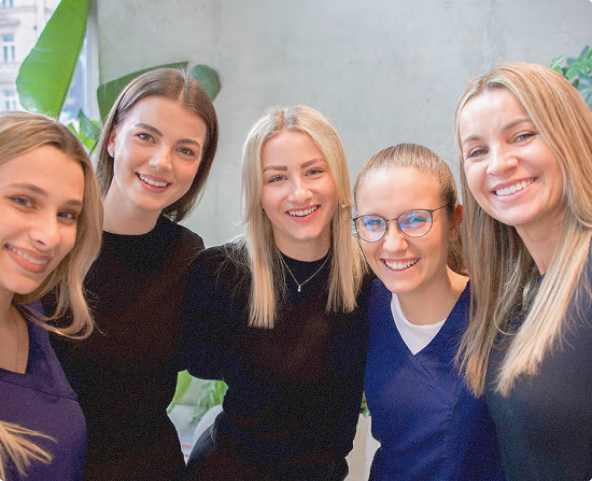 Pět usmívajících se mladých žen pózuje společně uvnitř místnosti s rostlinami na pozadí.