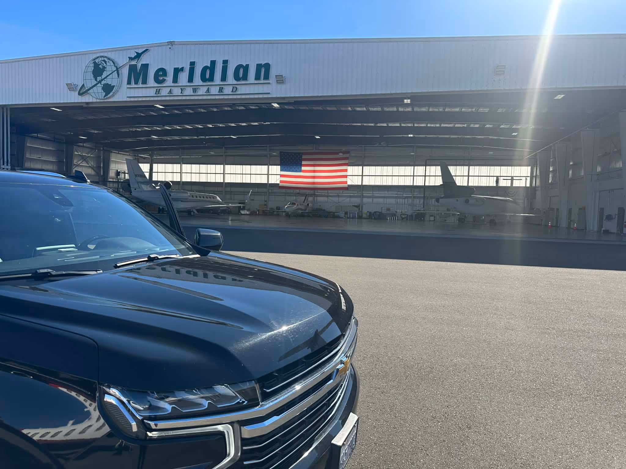 Front of a black Chevrolet vehicle parked outside a Meridian Hayward airplane hangar with private jets and an American flag inside.