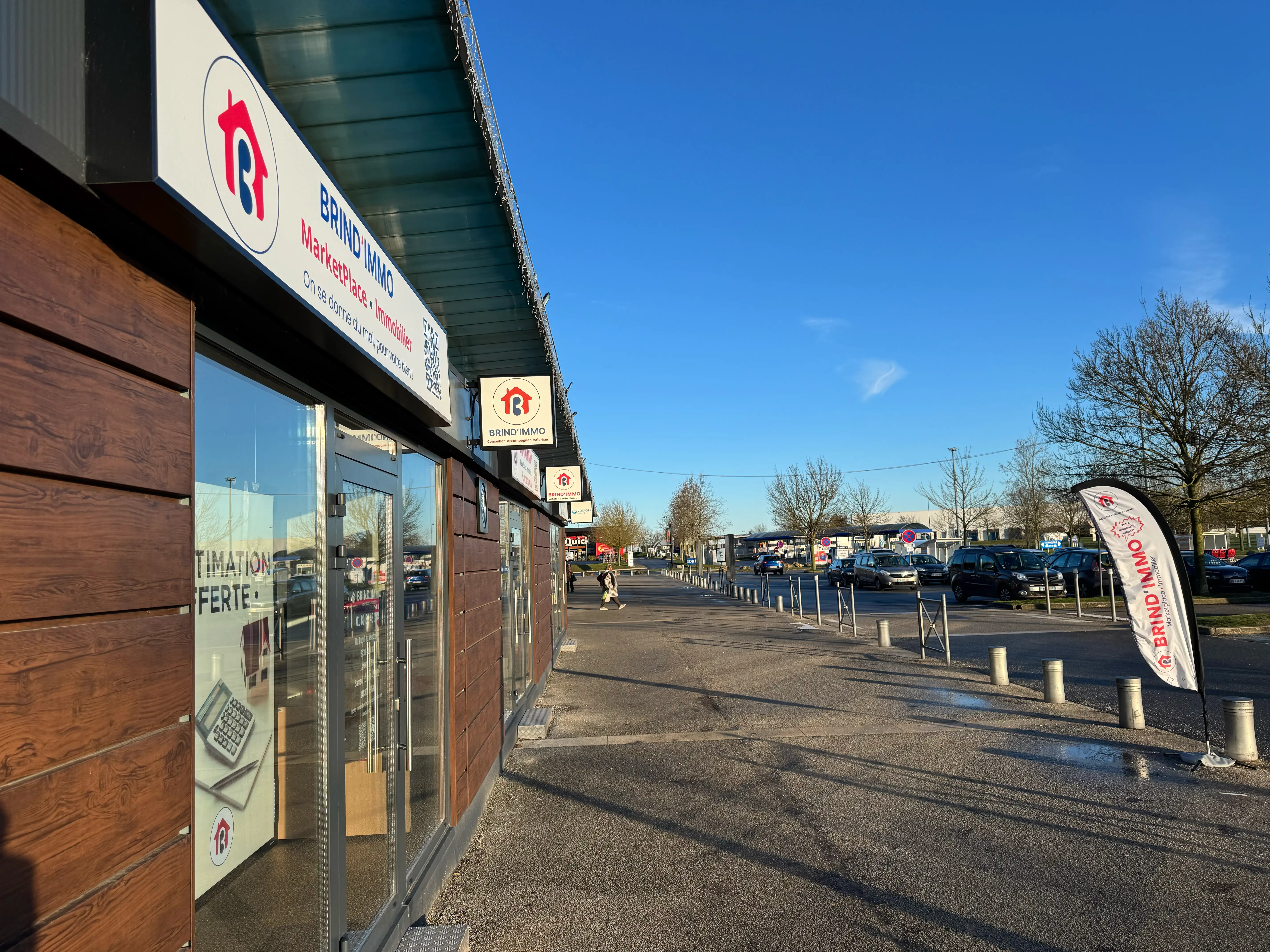 Façade d'agence immobilière BRIND'IMMO avec signalétique externe sous un ciel bleu dégagé et parking à côté.