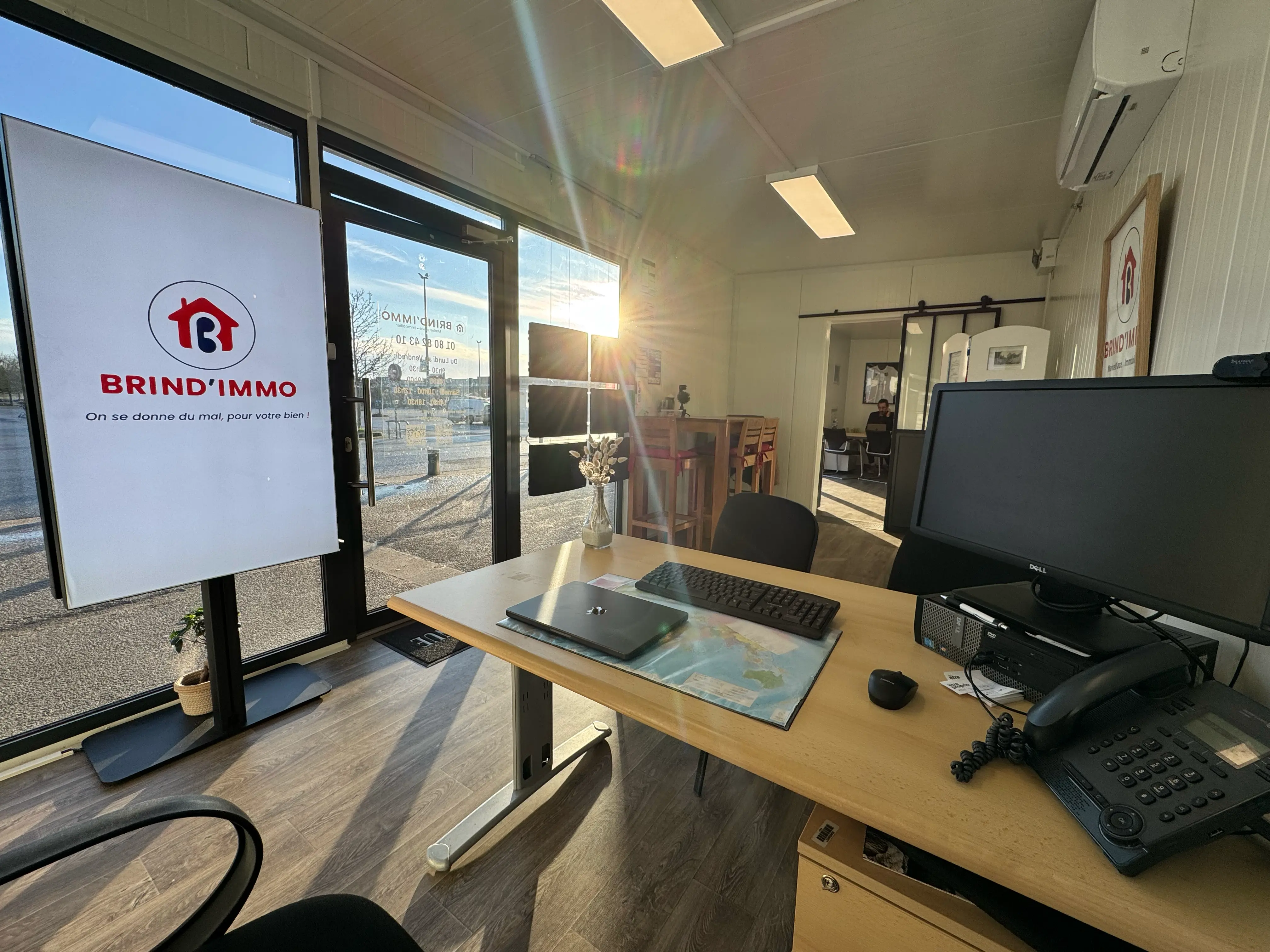 Bureau lumineux avec ordinateur, clavier, téléphone, ordinateur portable fermé, et vase avec fleurs séchées, doté d'une grande vitrine avec logo Brind'Immo en rouge.
