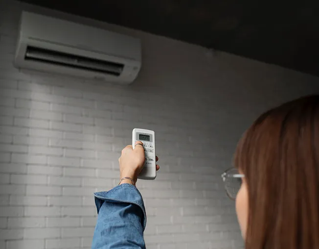 Person with glasses wearing blue denim jacket pointing a remote control towards a wall-mounted air conditioner.