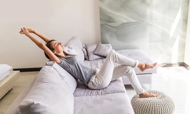 Woman in casual clothes stretching and relaxing on a light gray sofa in a bright living room.