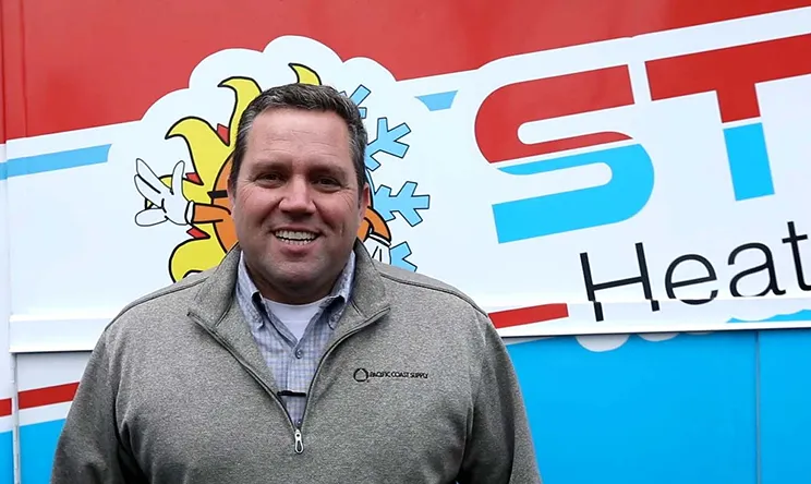 Smiling man in gray zip-up jacket standing in front of a colorful heating and cooling company sign.
