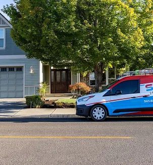 White and blue service van parked on a street next to a house entrance shaded by a large leafy green tree.