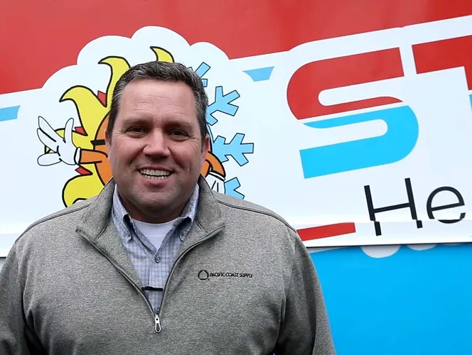 Smiling man wearing a gray zip-up jacket standing in front of a colorful sign with sun and snowflake graphics.