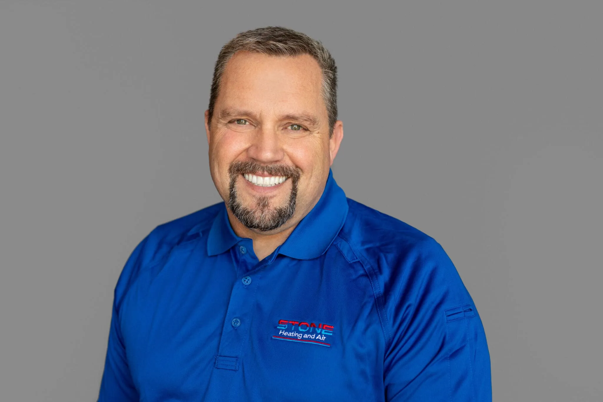Smiling man wearing a blue shirt with Stone Heating and Air logo on a gray background.