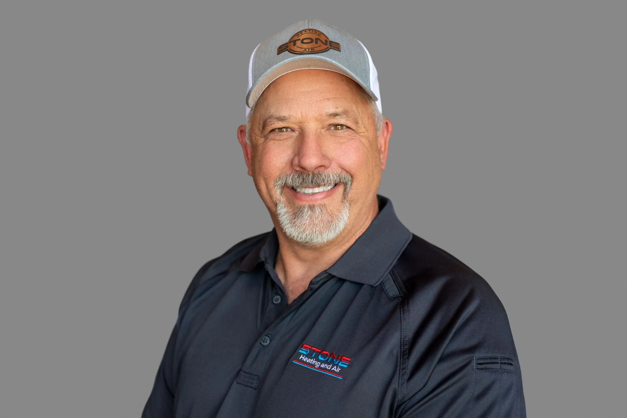 Smiling middle-aged man with gray beard wearing a Stone Heating and Air cap and polo shirt on a gray background.