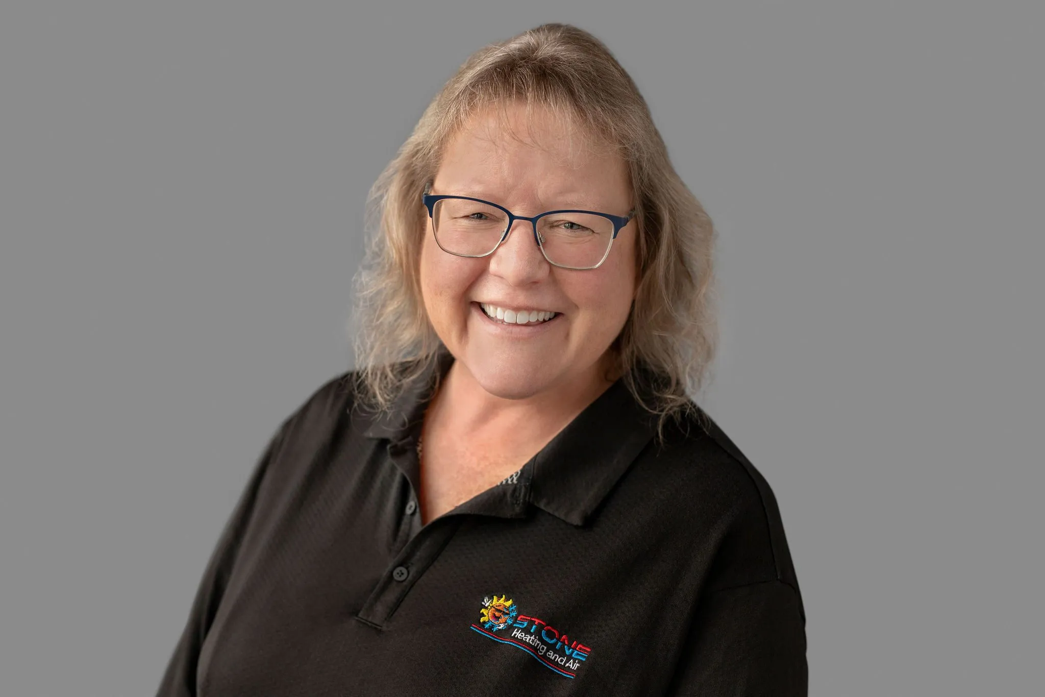 Smiling woman with glasses wearing a black polo shirt with Stone Heating and Air logo on a gray background.