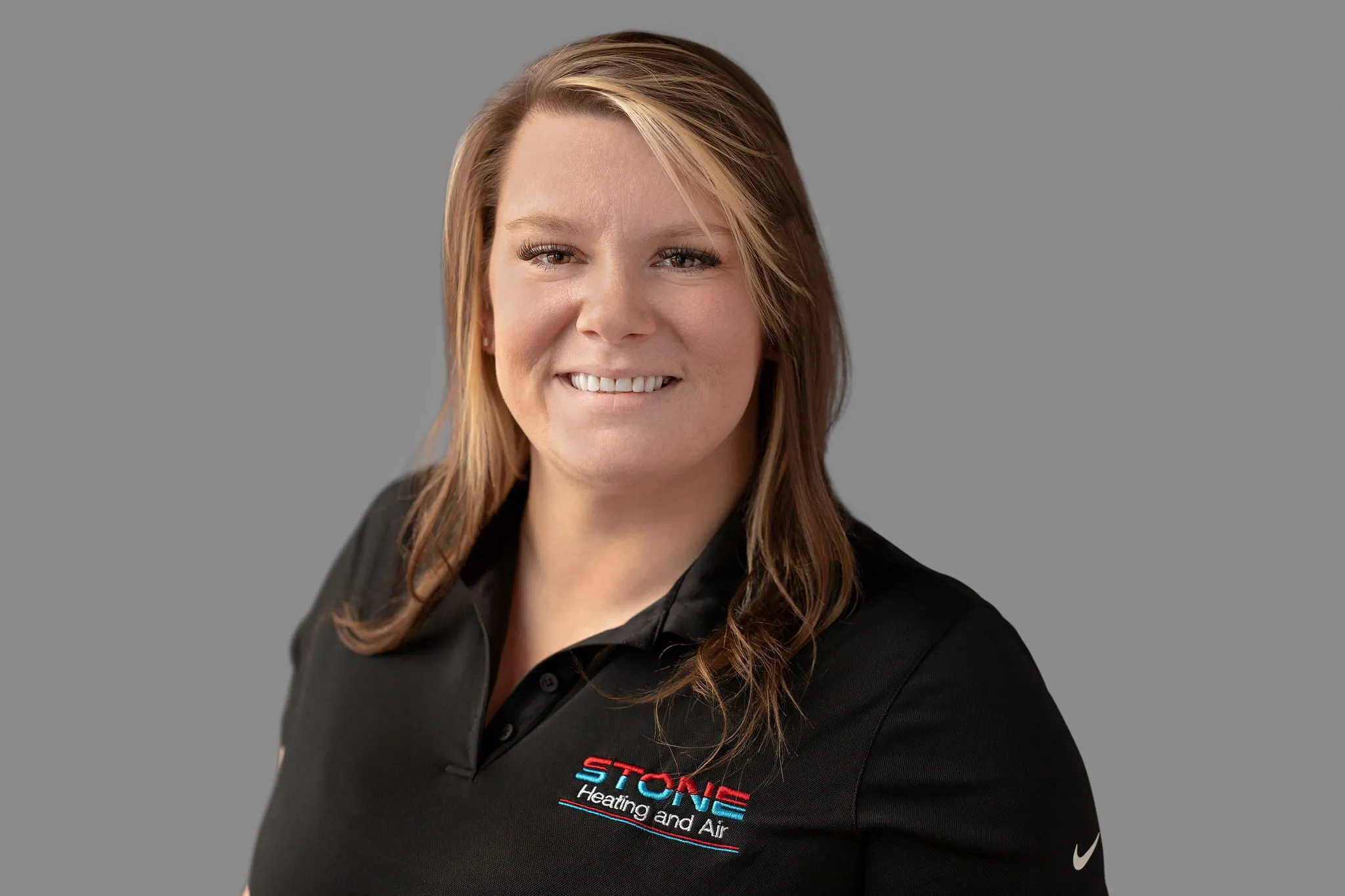 Smiling woman with light brown hair wearing a black Stone Heating and Air polo shirt against a gray background.
