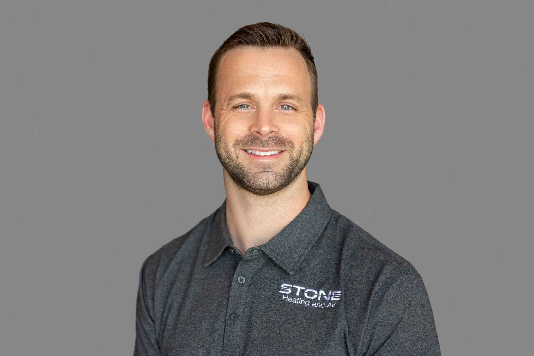 Smiling man wearing a dark gray polo shirt with 'STONE Heating and Air' logo on a gray background.