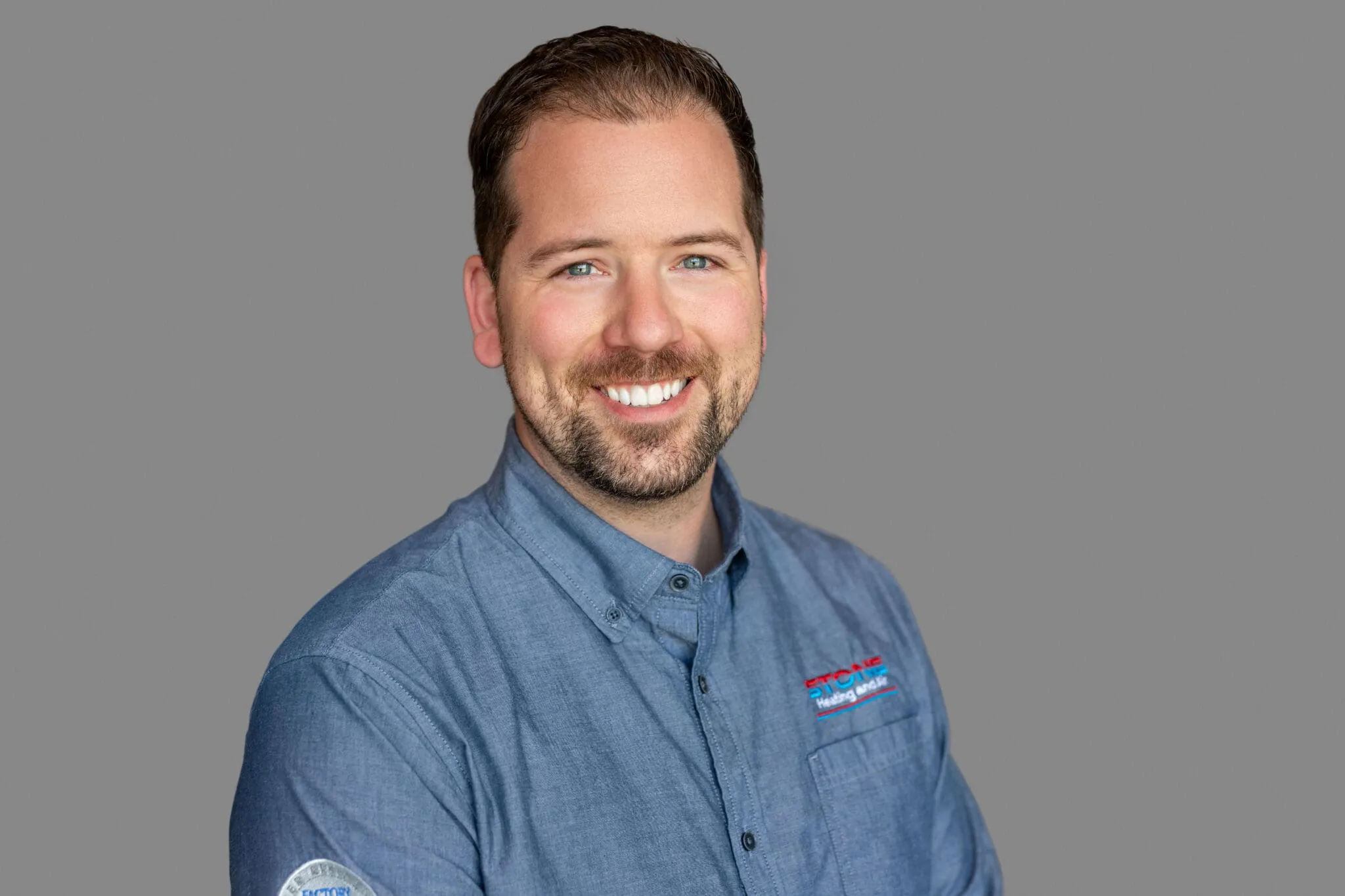 Smiling man with short brown hair and beard wearing a blue button-up shirt with company logos on chest and sleeve, against a plain gray background.