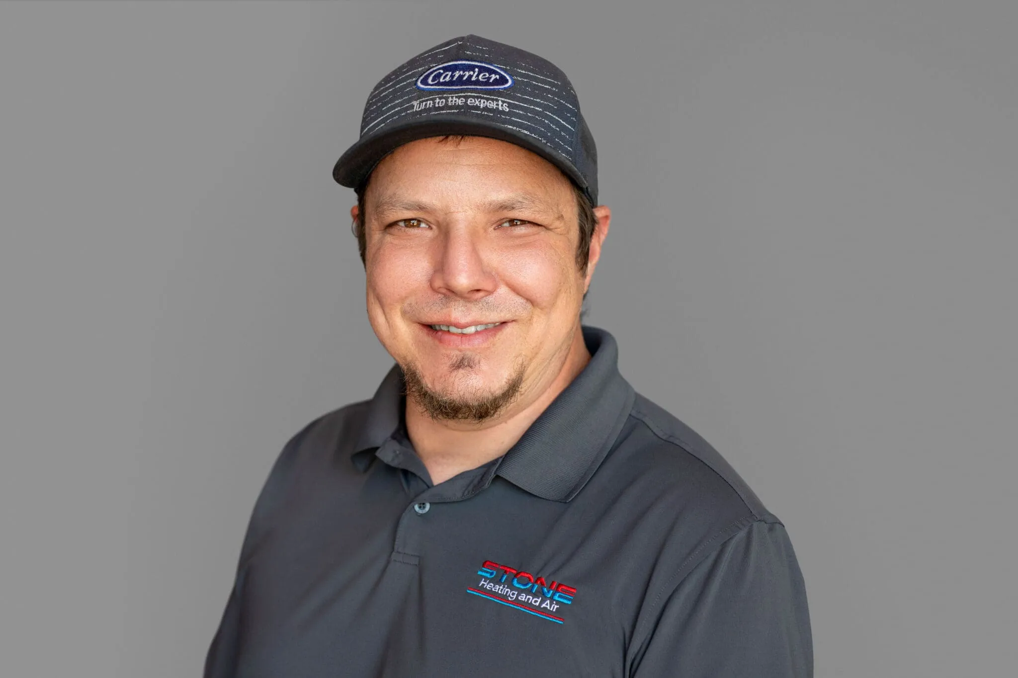 Smiling man wearing a Carrier cap and gray polo shirt with Stone Heating and Air logo on a gray background.
