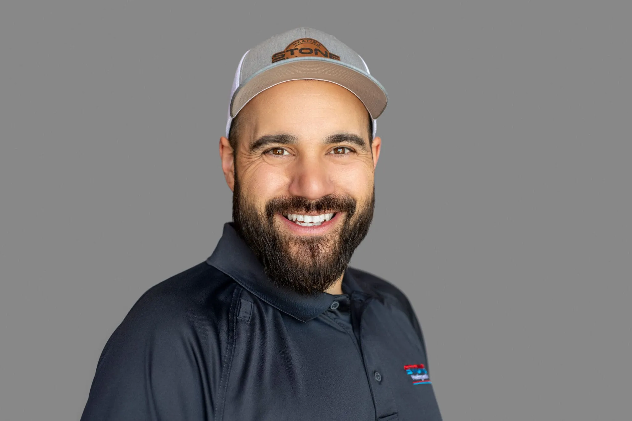 Smiling man with beard wearing a gray cap and dark polo shirt against a plain gray background.