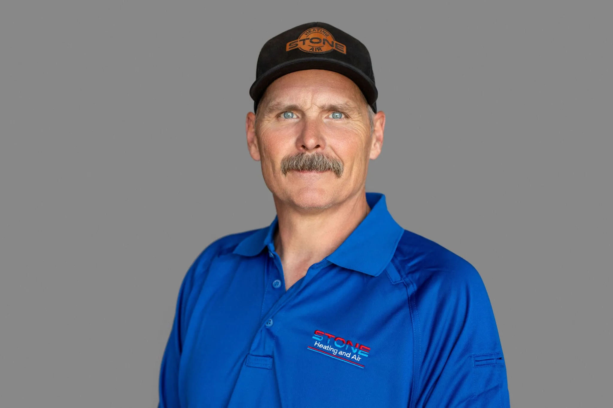 Portrait of a man with a mustache wearing a black Stone Heating and Air cap and a blue Stone Heating and Air polo shirt.