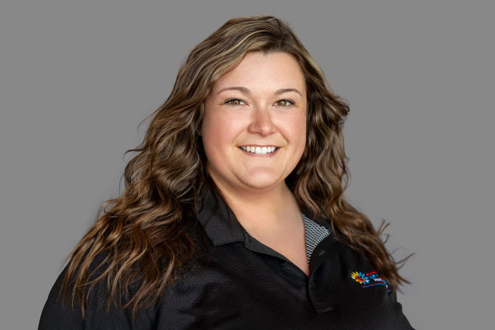 Smiling woman with long wavy brown hair wearing a black collared shirt against a gray background.