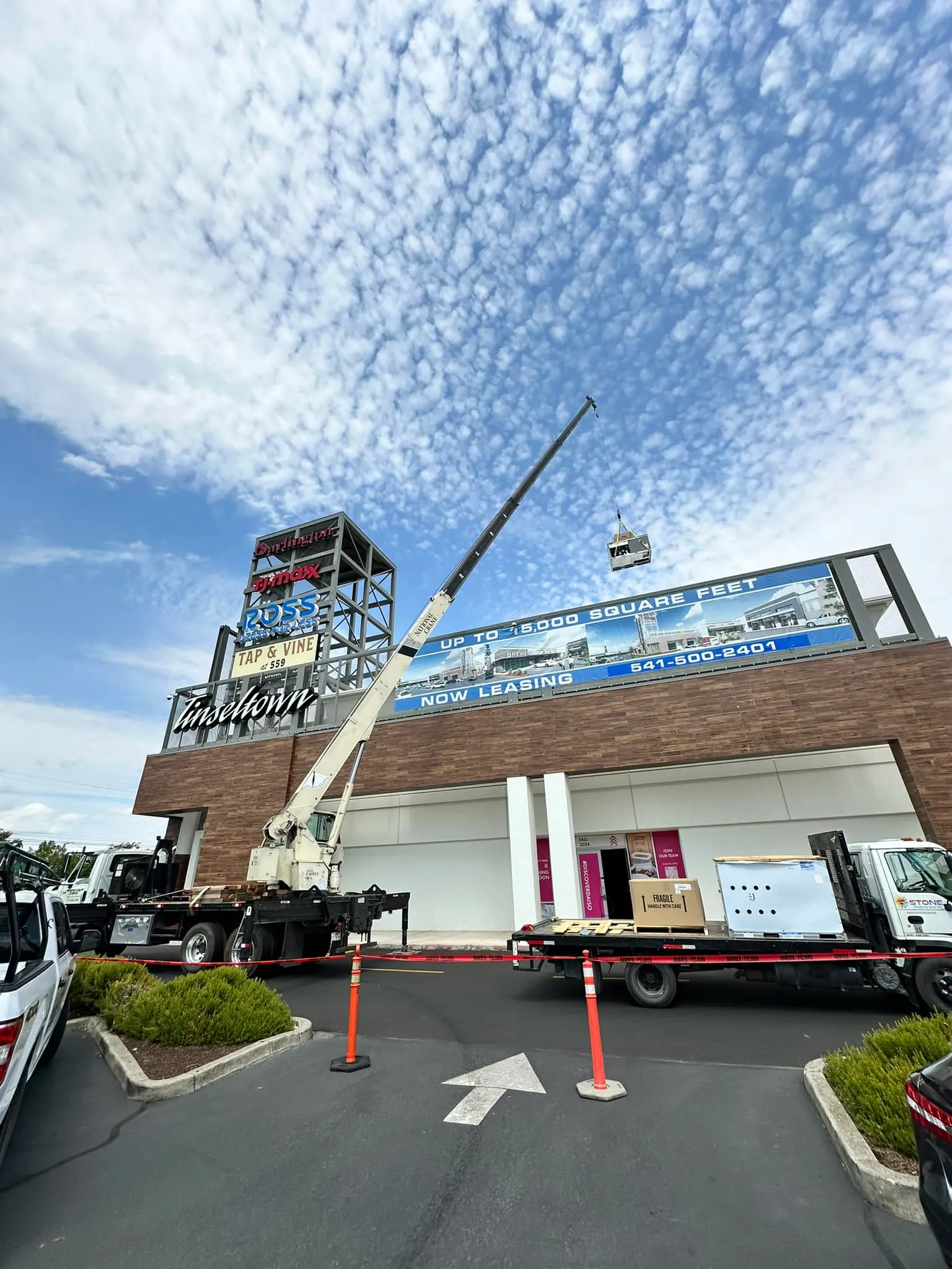 Crane lifting an air conditioning unit onto a building with retail signs under a cloudy blue sky.