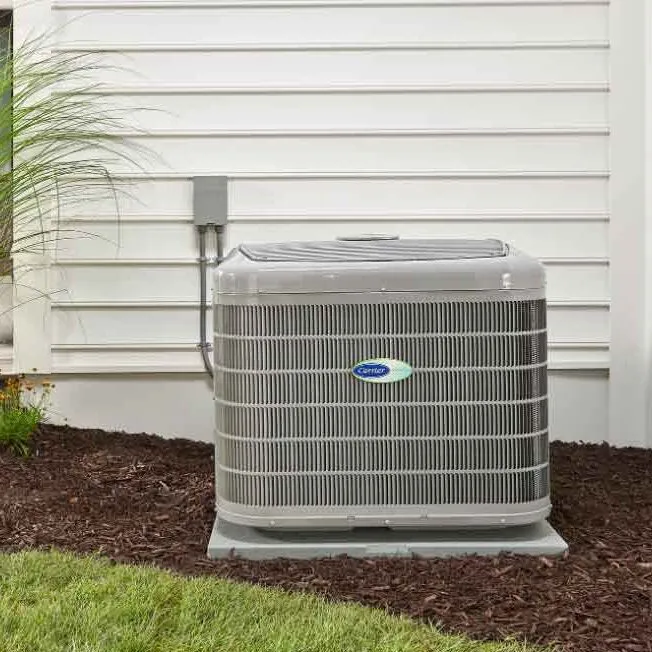 Carrier brand outdoor air conditioning unit installed next to a white siding house with grass and mulch around it.