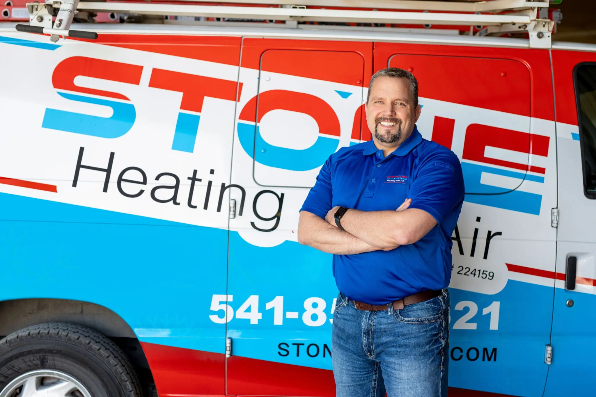 Smiling man in blue polo and jeans stands with arms crossed in front of a red, white, and blue heating and air service van.