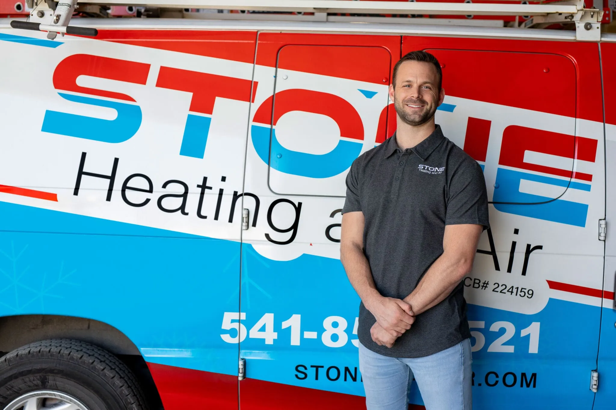 Smiling man in dark gray polo and light jeans standing in front of a Stone Heating and Air service van.