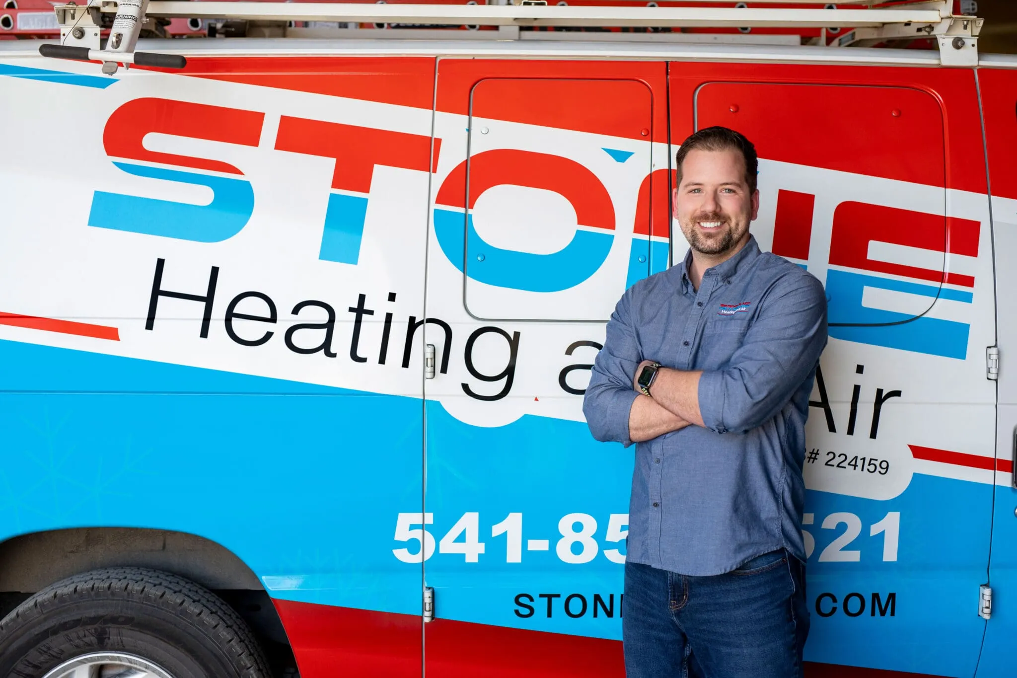 Smiling man with crossed arms standing in front of a Stone Heating and Air service van.