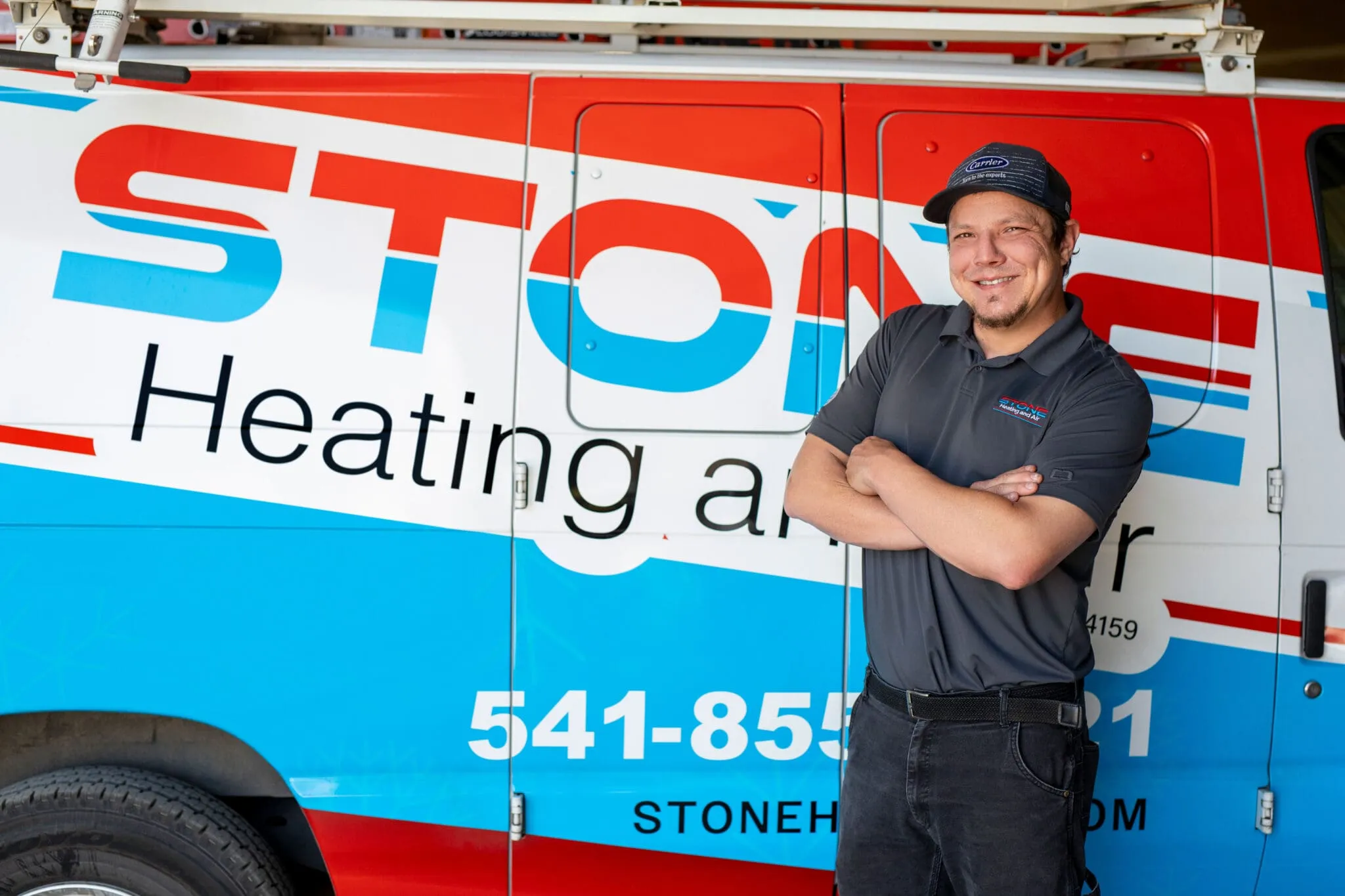Smiling man in dark uniform and cap standing with arms crossed in front of a colorful Stone Heating and Air service van.