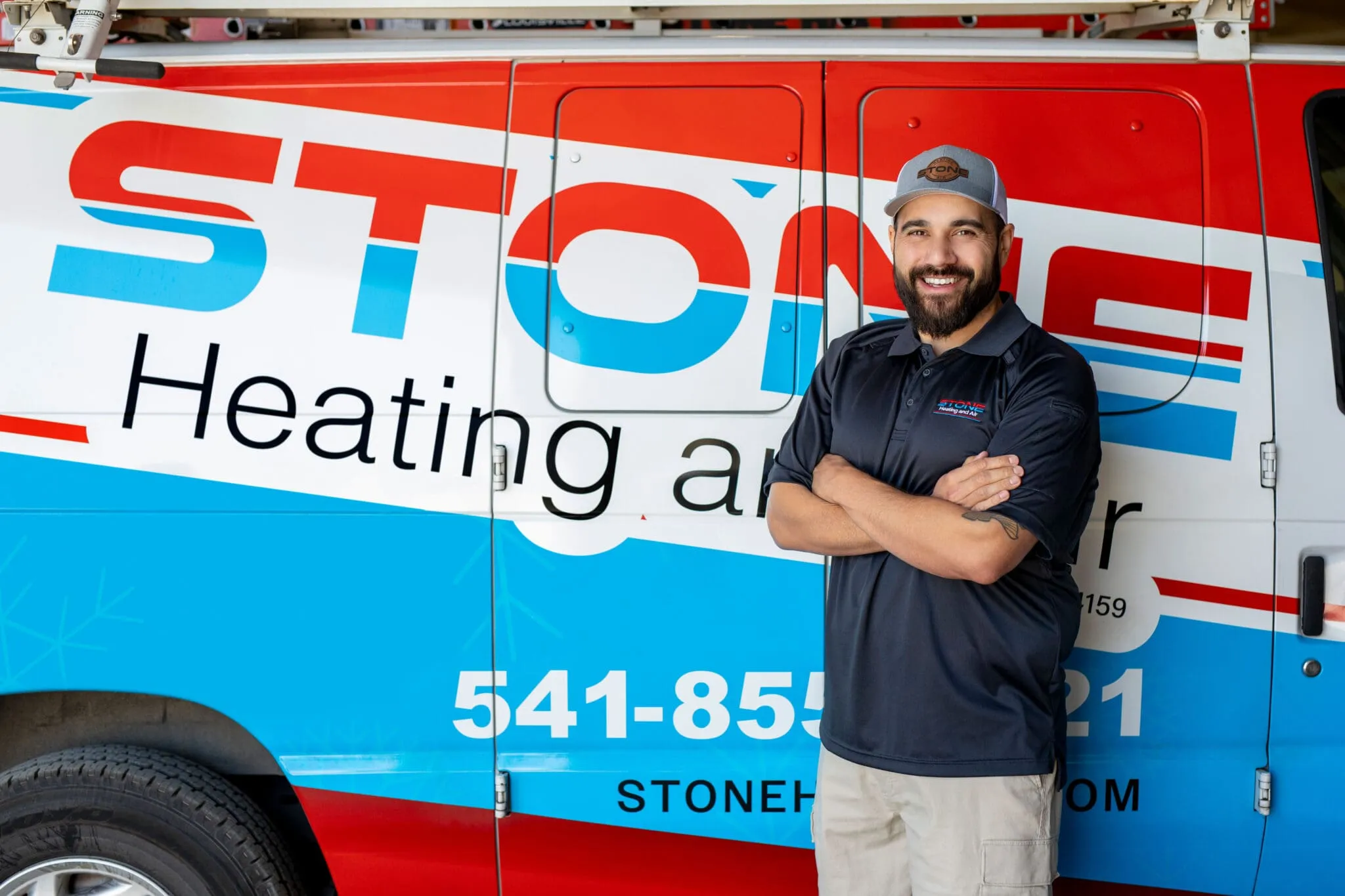Smiling man with a beard wearing a Stone Heating and Air cap and shirt standing with arms crossed in front of a Stone Heating and Air service van.