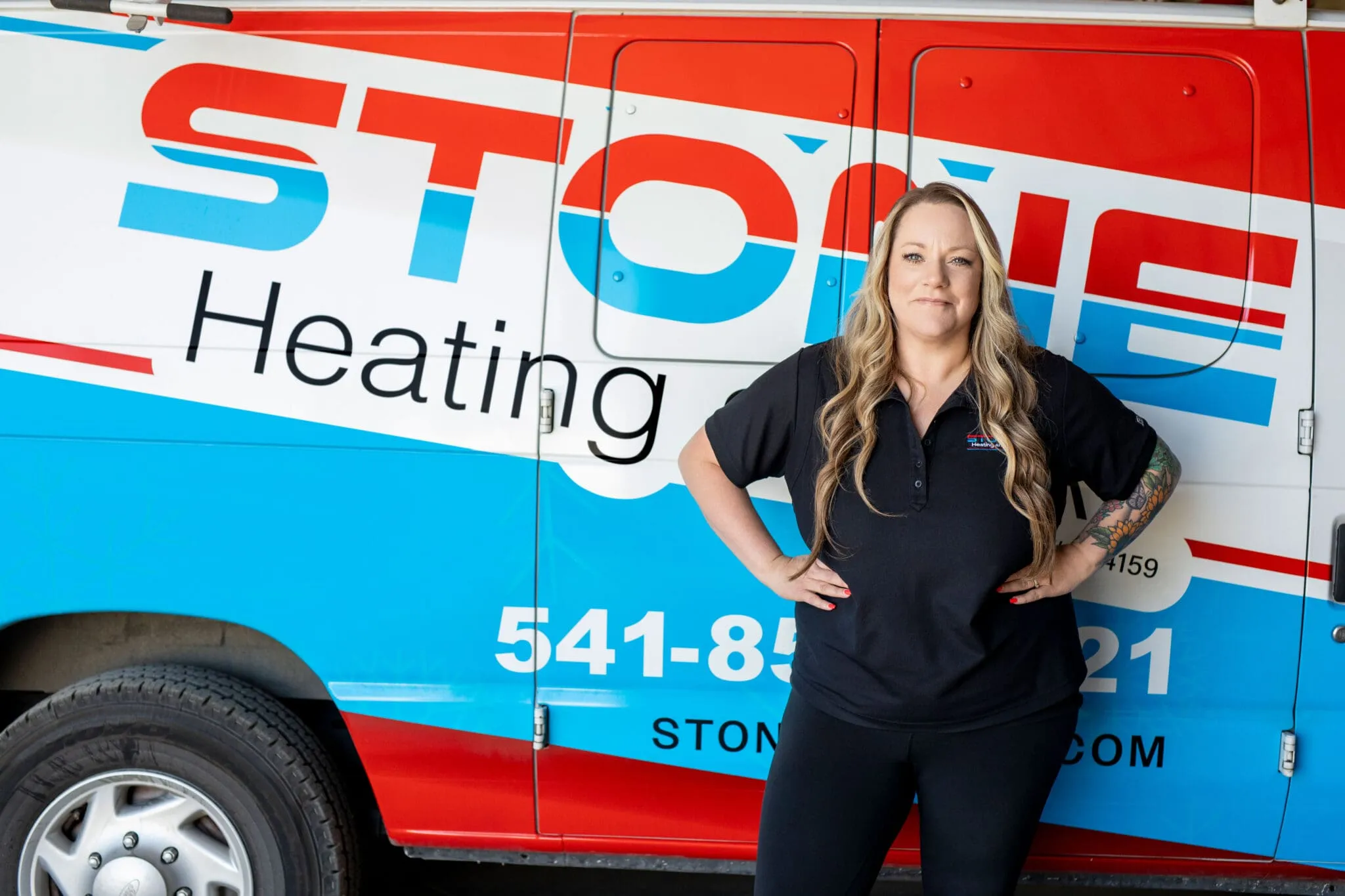 Woman with long blonde hair and a tattoo on her left arm stands with hands on hips in front of a red, white, and blue service van with "Stone Heating" branding.
