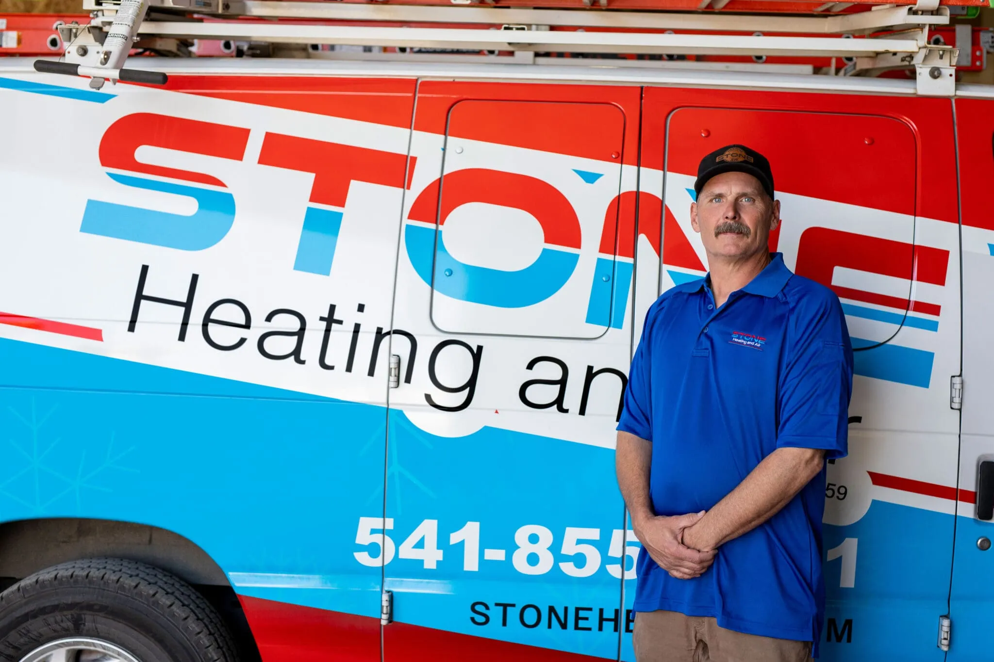 Man in a blue polo and black cap standing in front of a Stone Heating and Air service van.